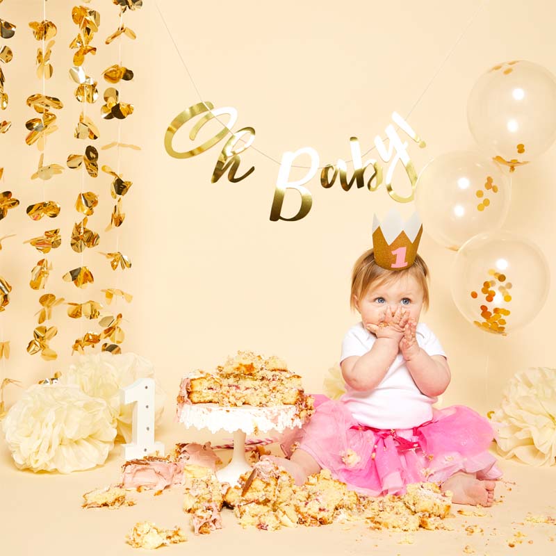 Baby celebrating a first birthday with a cake, gold Oh Baby bunting and confetti balloons.