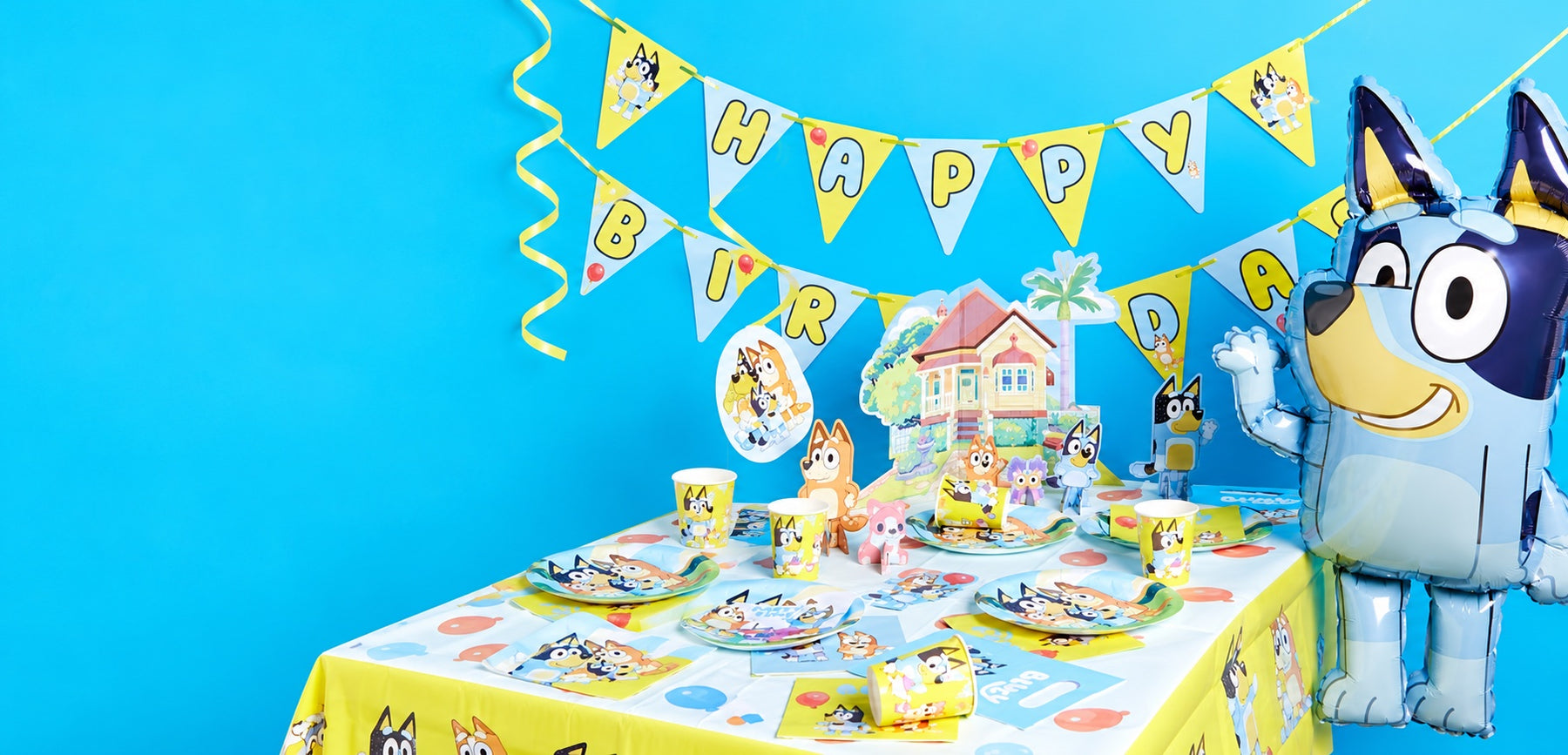 Bluey birthday party with a character balloon, yellow bunting and a table of Bluey plates, cups and decorations.