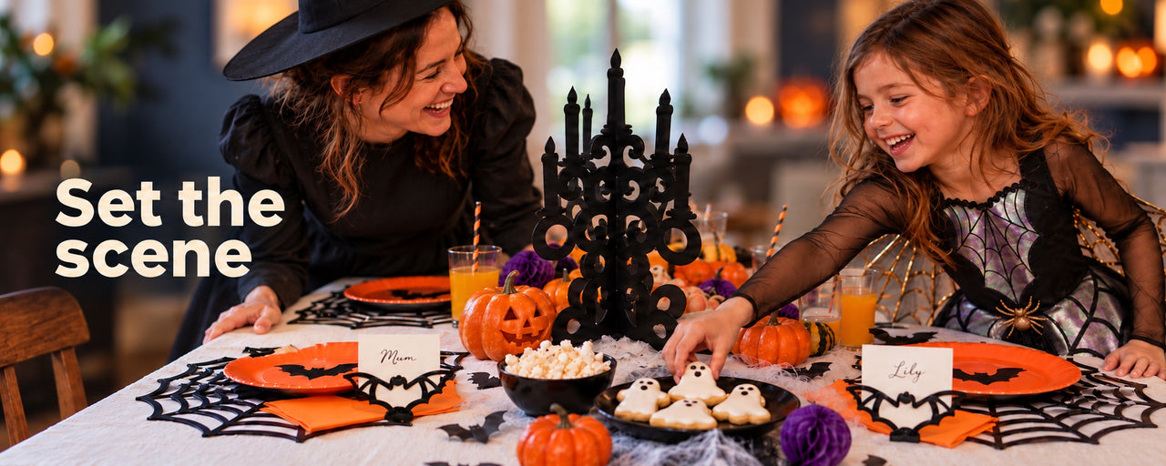Set the scene: a mother in a Wicked Witch costume and a child in an Iridescent Spider Girl costume laughing over a Halloween table with a candelabra, bat place cards, spiderweb placemats and cobwebs.