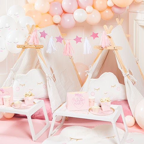 Two kids' sleepover tents set up for a sleepover party with pink blankets, cloud cushions, star-shaped bunting, a pastel balloon arch and bed trays with sleepover snacks placed on them