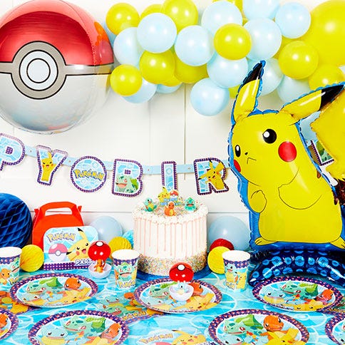 A Pokémon-themed party with a Pikachu balloon to the right and a Pokéball to the left joined together by a blue and yellow balloon arch and a Pokémon birthday banner balloon. There is a table with with Pokémon party tableware below, as well as a cake.