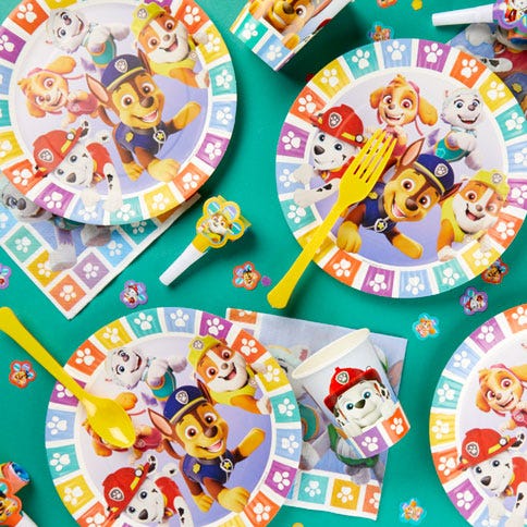 The picture shows a Paw Patrol party with a giant Chase balloon and a primary colour balloon arch. There is a table along the bottom with Paw Patrol paper plates and tableware, a cake and character masks.